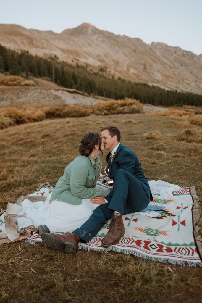 A bride and a groom sit on an boho blanket during their romantic sunset picnic at sunset during their Colorado elopement.