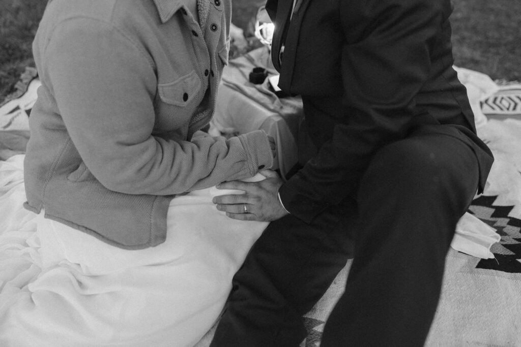 Black and white photo of the groom resting his hand on his new bride's leg during their romantic sunset picnic.