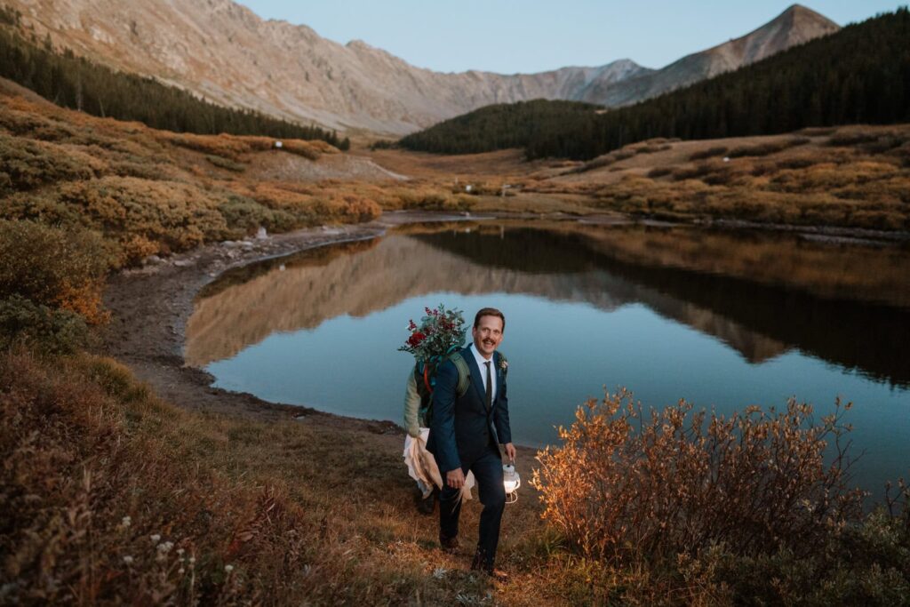 The groom poses for a photo with his backpack and a lantern in front of a still alpine lake near Breckenridge, Colorado, just after sunset.