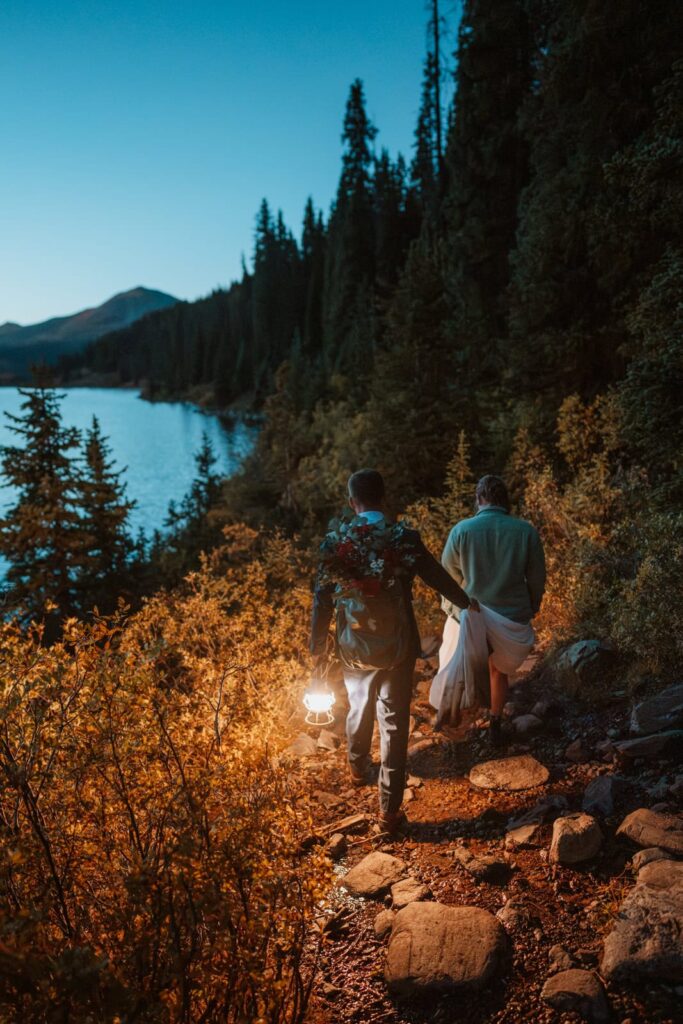 Bride and groom hike at dusk with bright lanterns along a hiking trail.