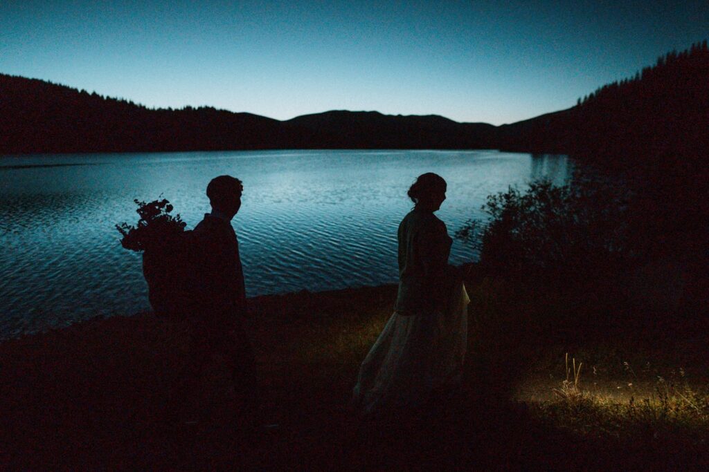A dramatic photo of a bride and groom walking in front of an alpine lake in Colorado after dark. You can only see their silhouettes.