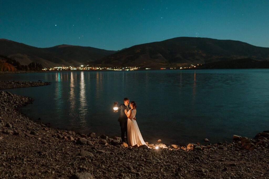 Newlyweds take star photos next to Dillon Reservoir in Breckenridge, Colorado. They are only lit by 2 small lanterns. The city lights reflect in the alpine lake in the background.