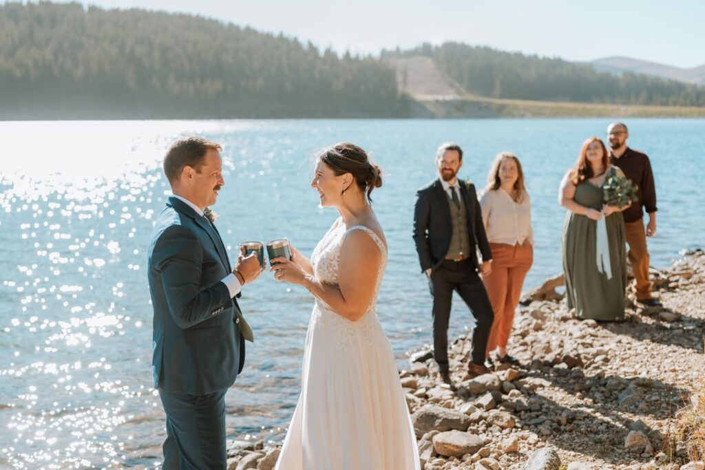 Bride and groom toast their coffee cups together in celebration of their small mountain wedding near Breckenridge. Their guests are in the background behind them and are smiling as they watch.