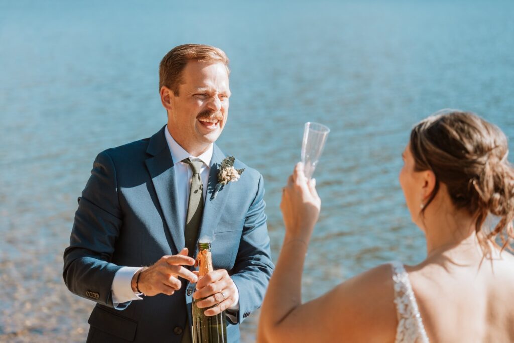 Bride holds up her champagne glass in celebration as the groom pops open the champagne.