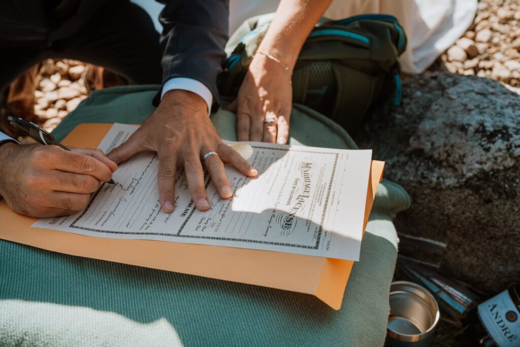 Closeup photo of the marriage license. The groom has his hand flat on the marriage license which shows off his wedding ring. He signs to make it official!