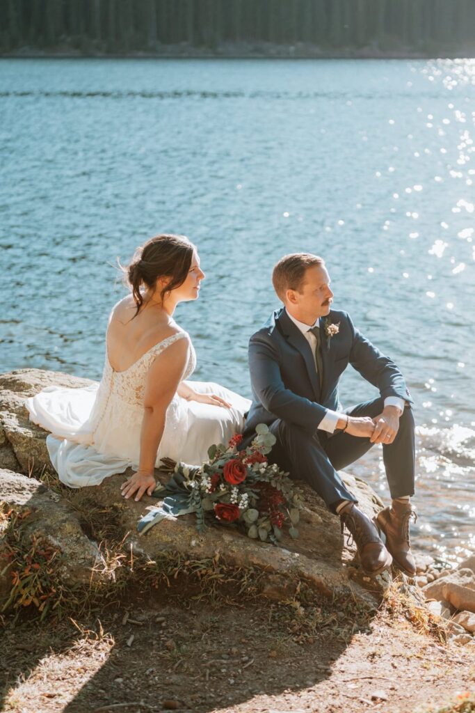 Bride and groom look off in the distance as they sit on a rock together next to an alpine lake.