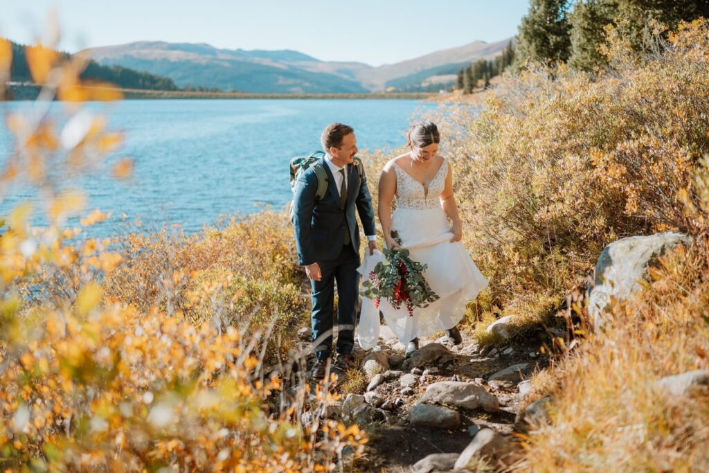 Bride and groom hiking on a rocky hiking trail with a large alpine lake in Colorado behind them. The foliage is yellow since it's mid-September.