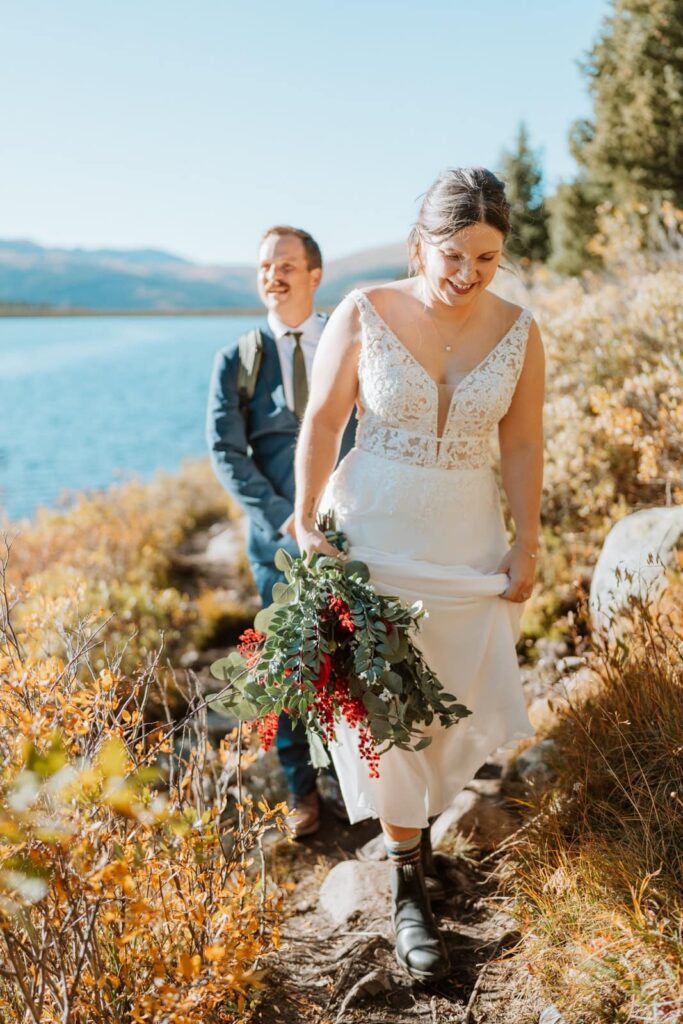 Bride lifts up the bottom of her dress as she holds her bouquet and walks along a hiking trail. Her groom is directly behind her with a smile on his face.
