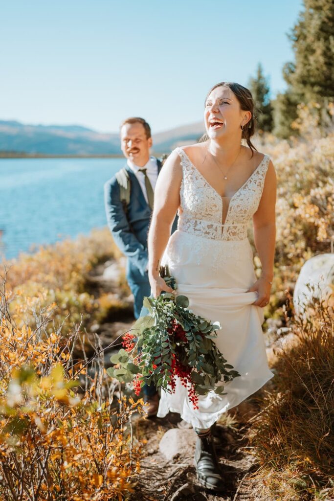 Bride laughs and smiles as she holds her dress and bouquet and hikes down a trail at Clinton Gulch reservoir in Colorado.
