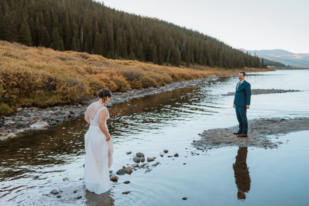 Bride stands on rocks in an alpine lake in Colorado as she gets ready to jump across the water to her groom.