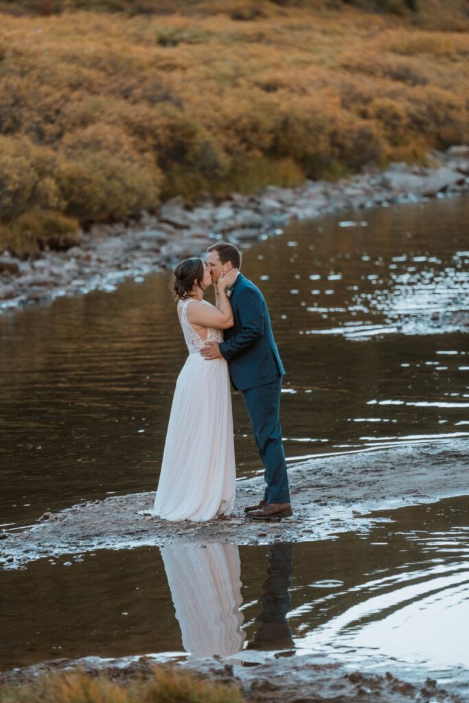 Bride and groom stand on a rock in an alpine lake at their small wedding in Colorado and share a kiss.