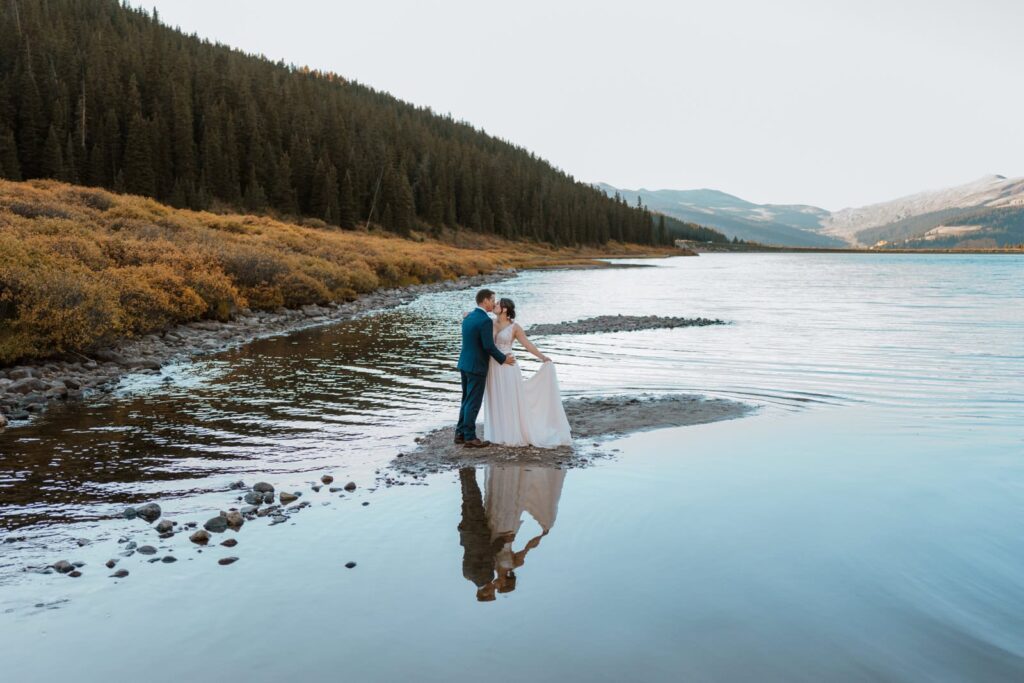 Bride fans out her dress as she kisses her groom while standing on a rock in an alpine lake for their small mountain wedding in Colorado. The water around them is so still that you can see their reflection.