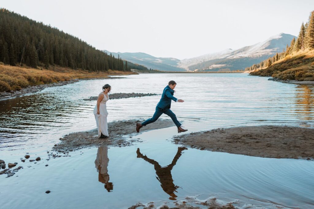 Groom hops from 1 rock to another rock that are completely surrounded by water. The bride is right behind him about to jump. Their reflections are cast in the alpine lake in front of them.