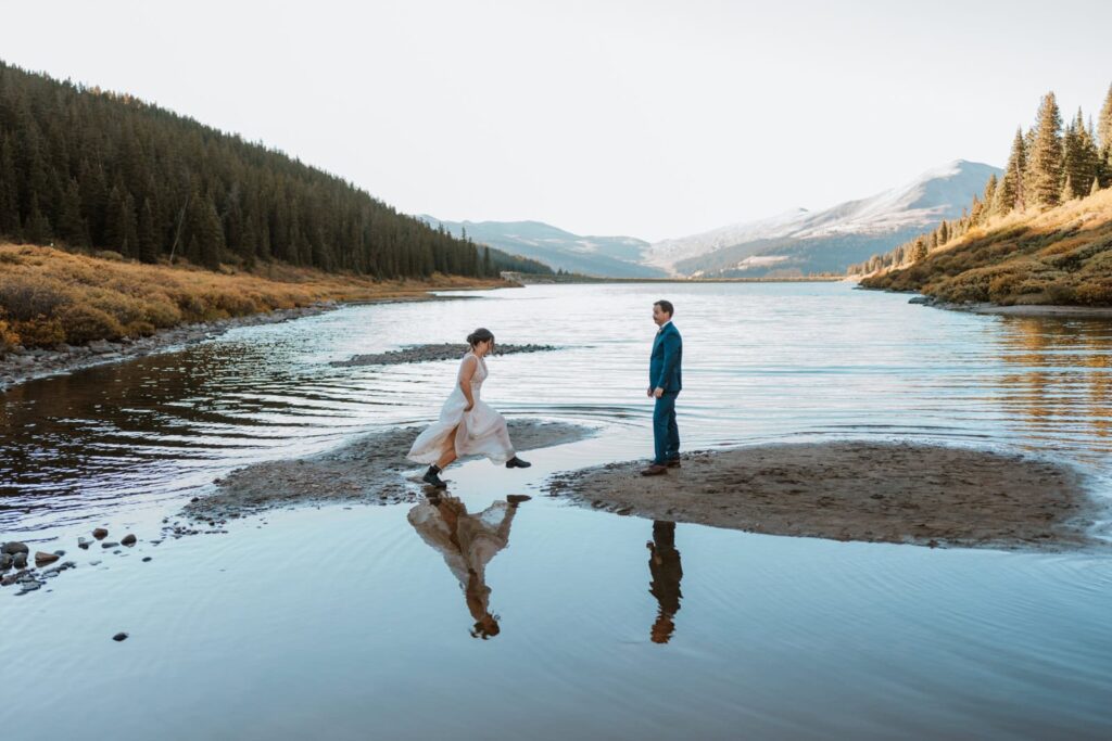 Bride jumps from one rock to another to her groom. They are surrounded by water and their reflections show in the alpine lake.
