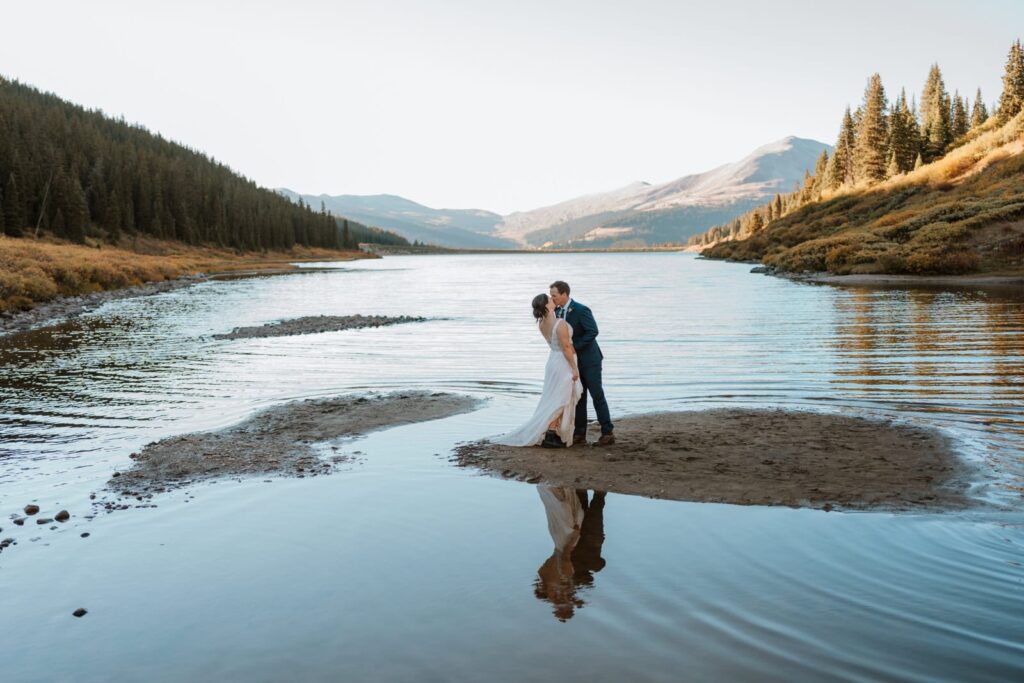 Bride and groom stand on a rock in an alpine lake in Colorado and share a kiss during their small mountain wedding in Colorado. Their reflections are cast back in the water in front of them.