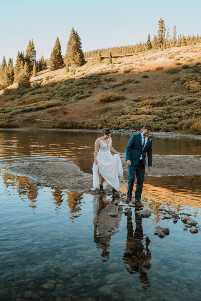 Bride and groom walk along the rocks in the water at sunset. Their reflections and the mountains are reflected back in the water in front of them.