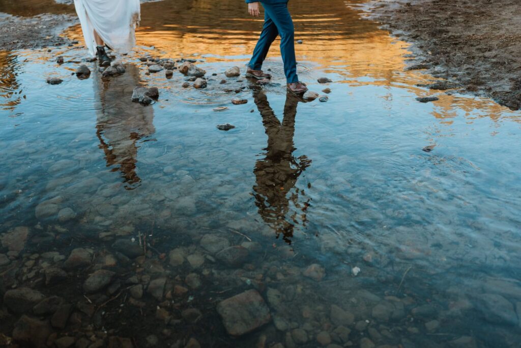 Detail photo shows the bride and grooms reflection in the alpine lake as they walk across the rocks.