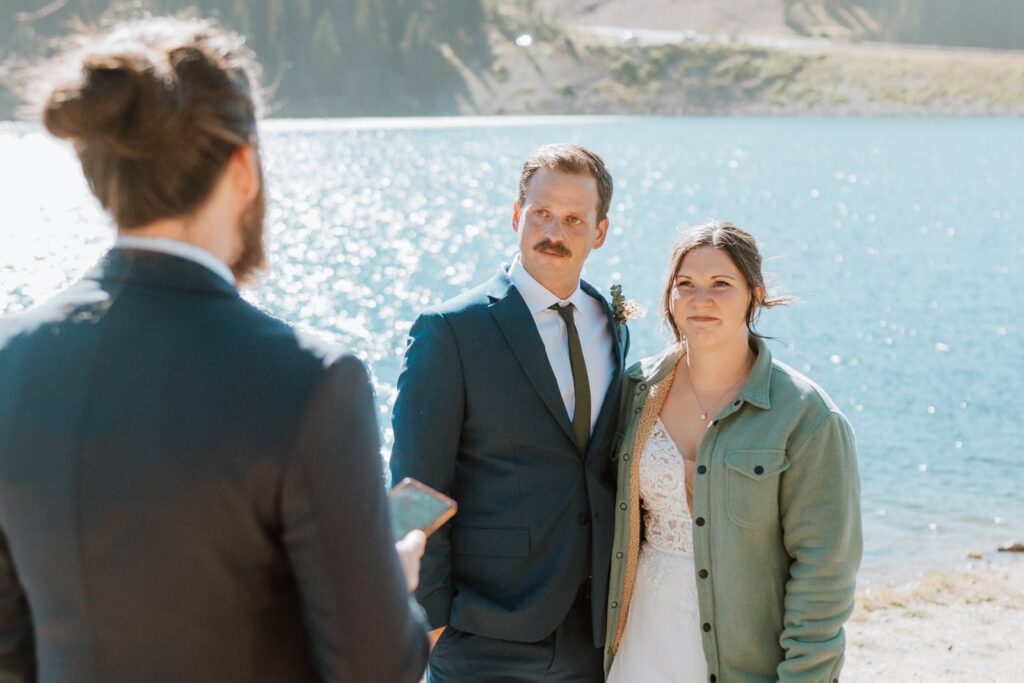 The bride and groom smile as the best man reads his speech. The best man is in the foreground and the wedding couple is photographed in front of him. Clinton gulch reservoir shines brightly behind them.