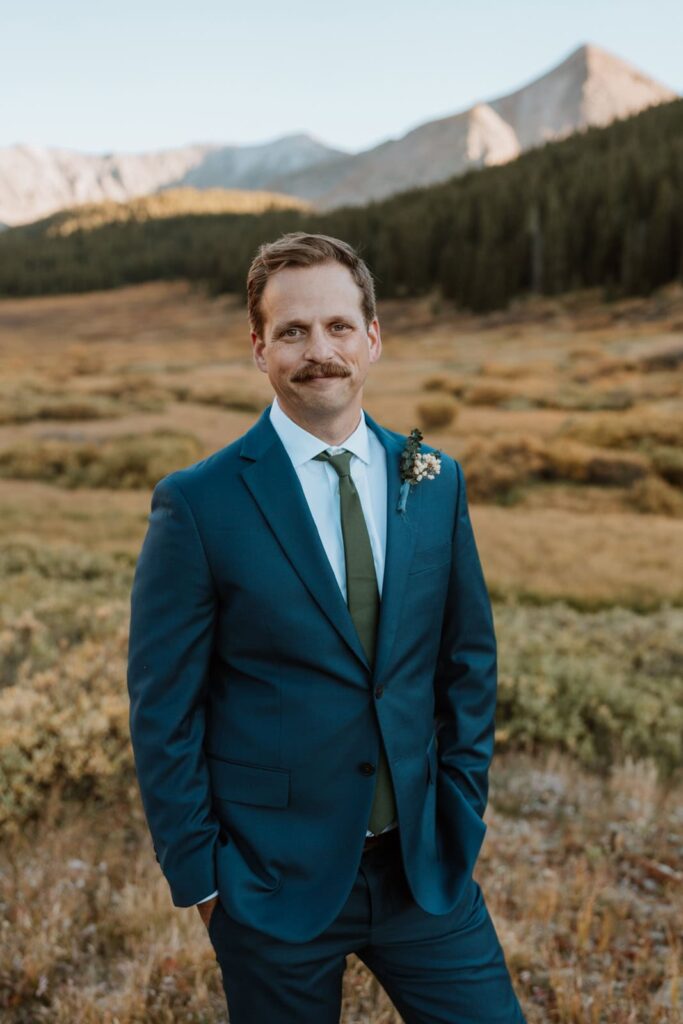 The groom poses for a photo in a navy blue suit for his small mountain wedding in Colorado. The mountains and fall foliage are vivid behind him.
