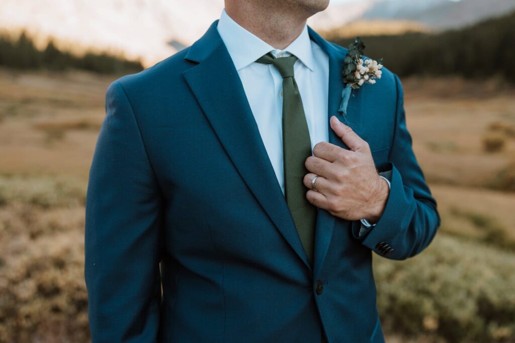 Closeup photo of the grooms blue suit and green tie. He holds onto the lapel to show off his wedding ring.