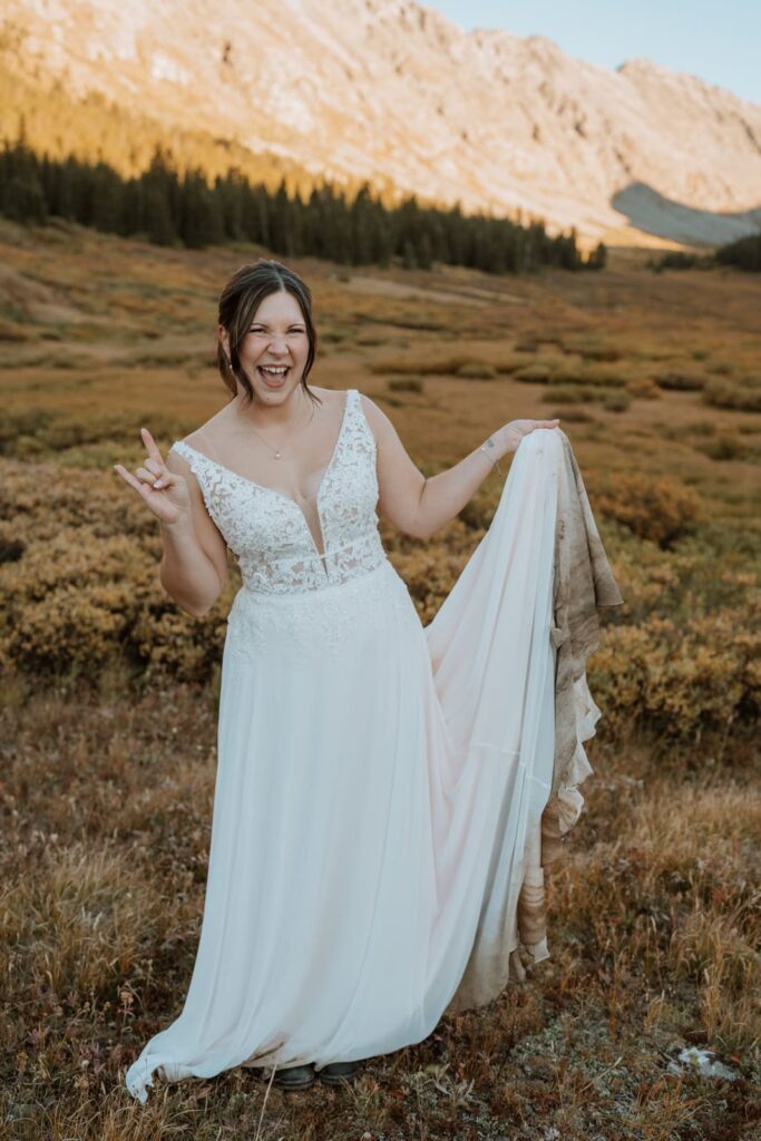 Bride holds up the end of her dirty wedding dress and throws up the 'rock on' symbol with her other hand for her small mountain wedding in Colorado.