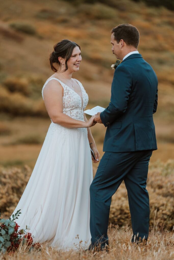 The bride has a huge smile on her face as her groom reads his vows to her and holds her hand.