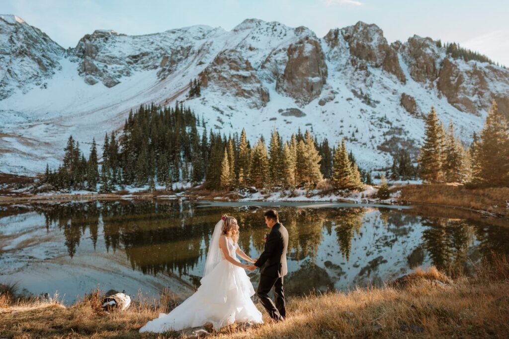 Bride and groom hold hands and prepare to read their wedding vows in front of a reflective alpine lake. The alpine lake is so still that it's reflecting the snow capped mountain peaks behind the couple. Telluride is a fantastic place to elope in Colorado if you want mountain views and luxury dining!