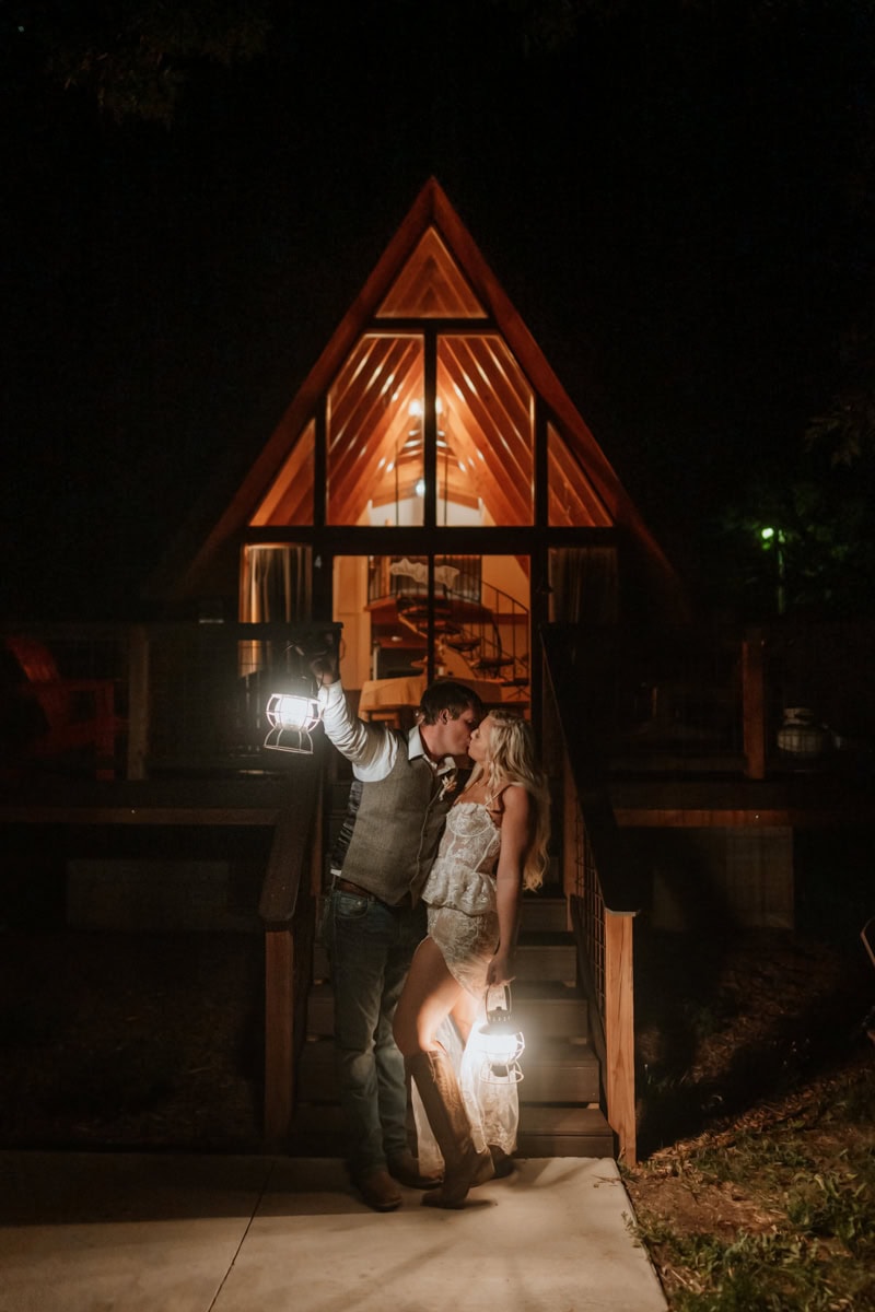 Bride and groom hold up lanterns and go in for a kiss. It's dark and they are standing in front of their a-frame cabin in Colorado holding lanterns.