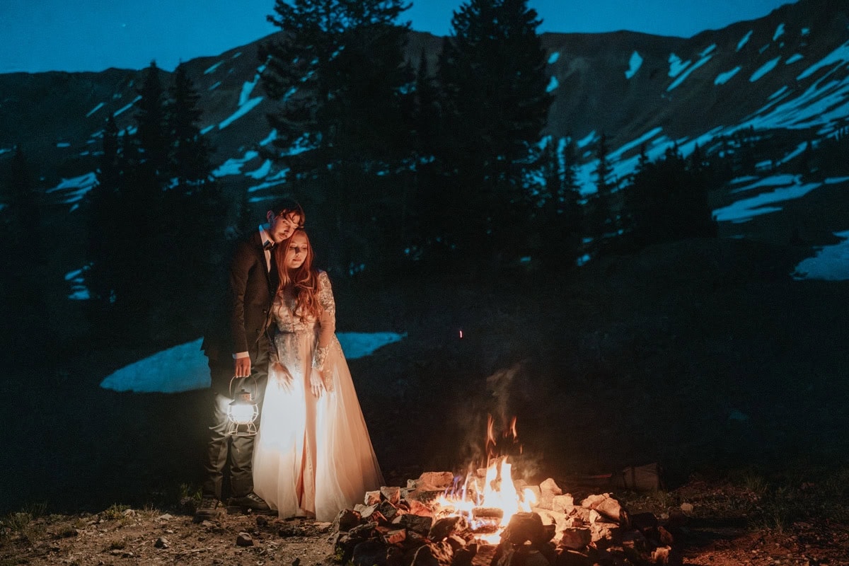 Bride and groom stand together holding a lantern while they look at a campfire burning during their Crested Butte elopement.
