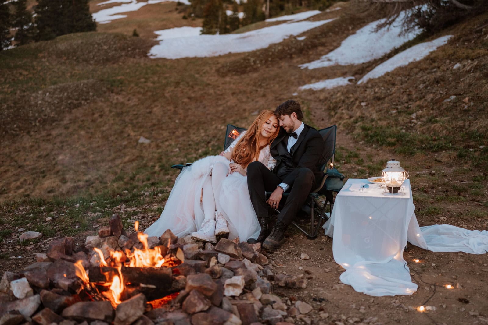 Couple relaxing by a campfire at sunset during a Crested Butte elopement, wrapped in blankets with mountain terrain and lingering snow patches in the background