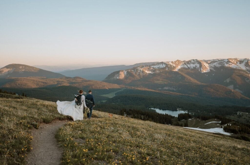Eloping couple walks down a hiking trail at sunset after reading their vows on top of a mountain for their Crested Butte eloepment. There are flowers all over around them and the sunset is lighting up a massive mountain range in the distance that still has small patches of snow on it.