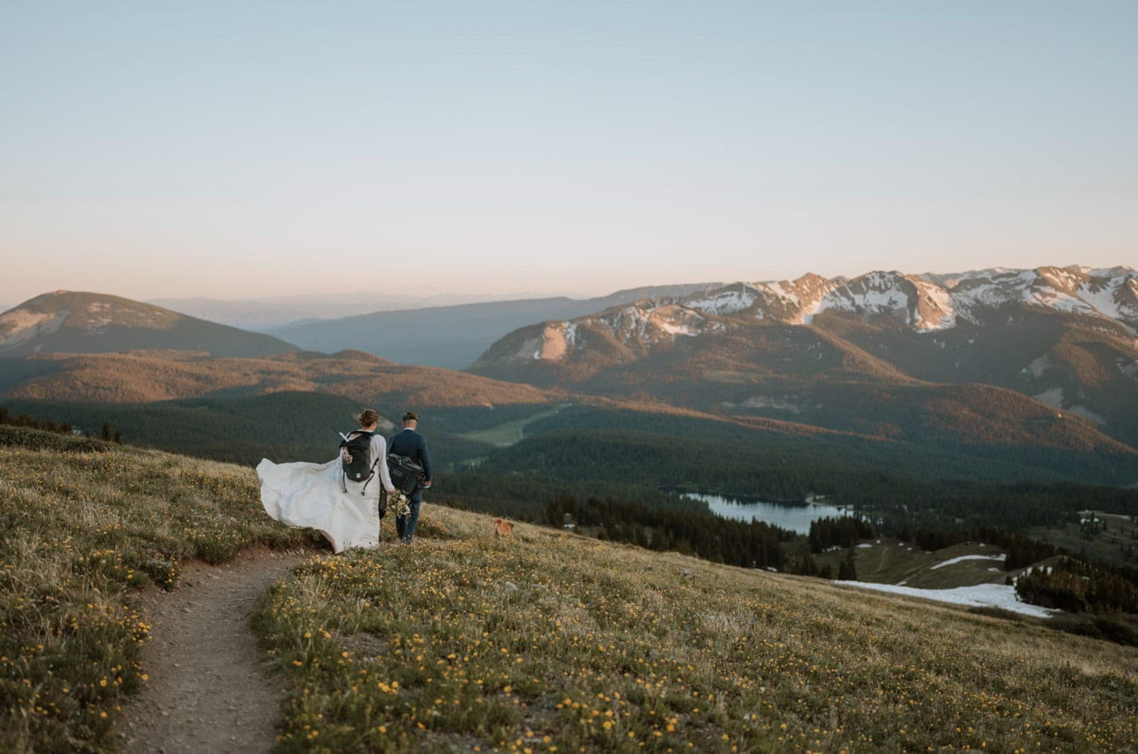 Eloping couple walks down a hiking trail at sunset after reading their vows on top of a mountain for their Crested Butte eloepment. There are flowers all over around them and the sunset is lighting up a massive mountain range in the distance that still has small patches of snow on it.