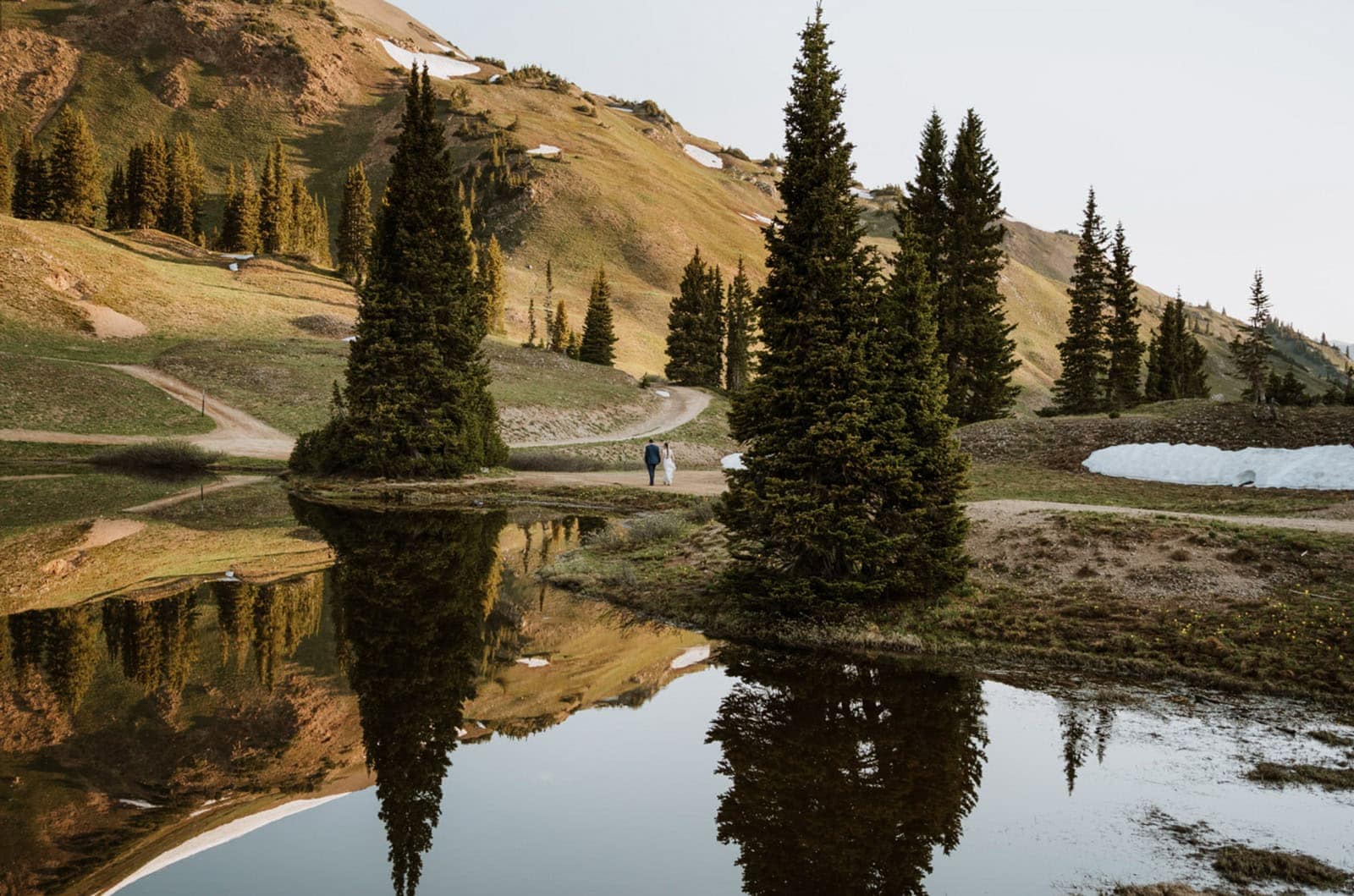 A newlywed couple walks at sunrise next to a reflective alpine lake in Crested Butte on a mountain pass. The pine trees are reflected back in the lake as they walk between them.