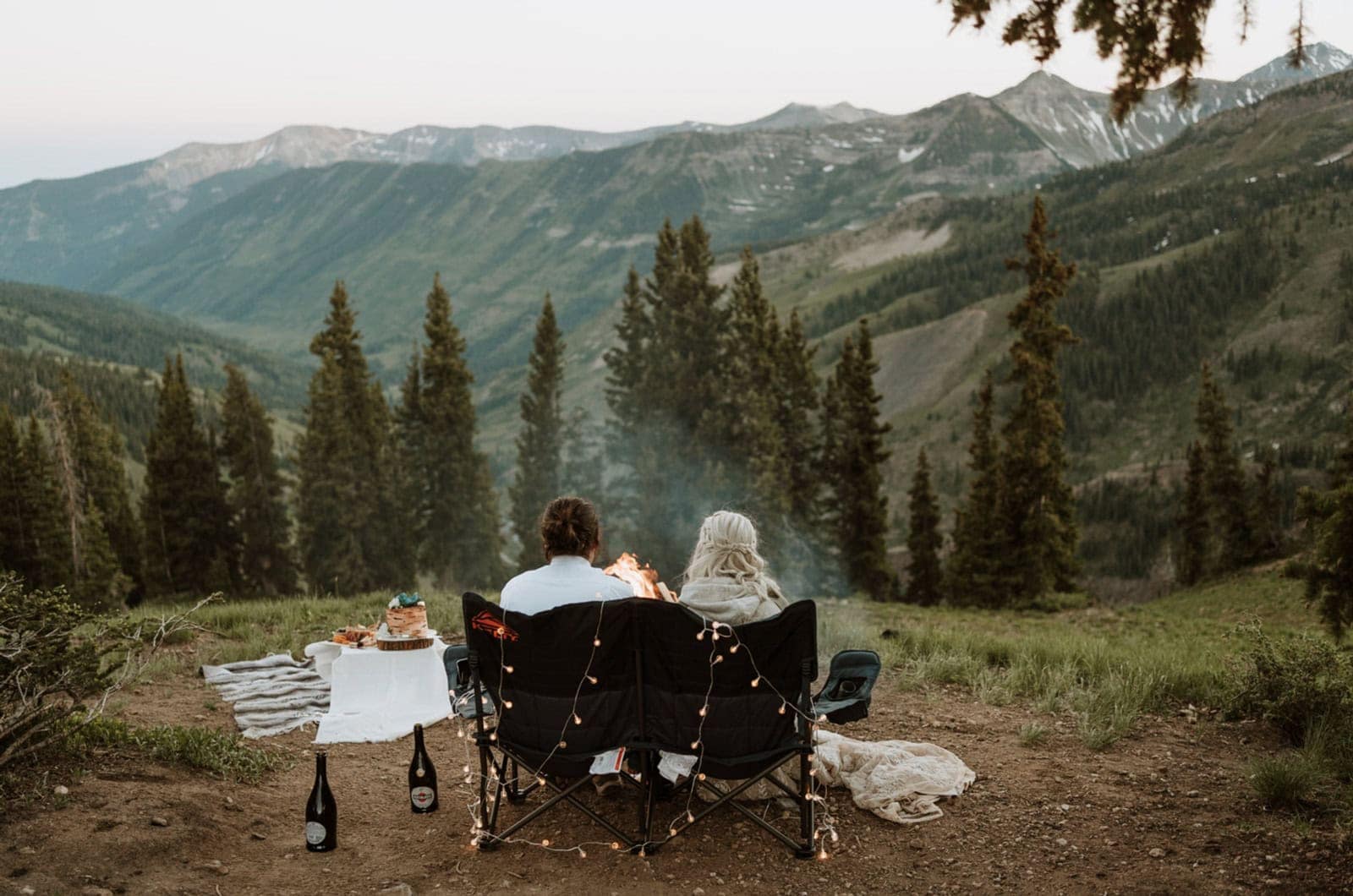 Bride and groom celebrate their Crested Butte elopement by sitting in a double loveseat Kelty campchair, drinking champagne, eating cake and enjoying a campfire with a view.