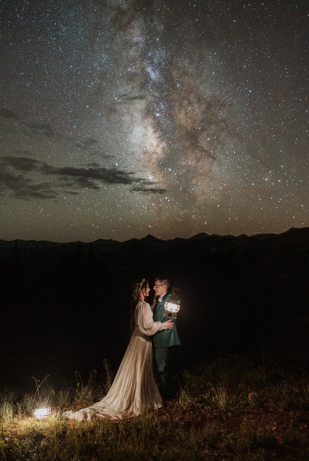 A wedding couple stands lit up by lanterns cuddling in the darkness after they eloped in Crested Butte. The milky way is bright in the sky behind them.