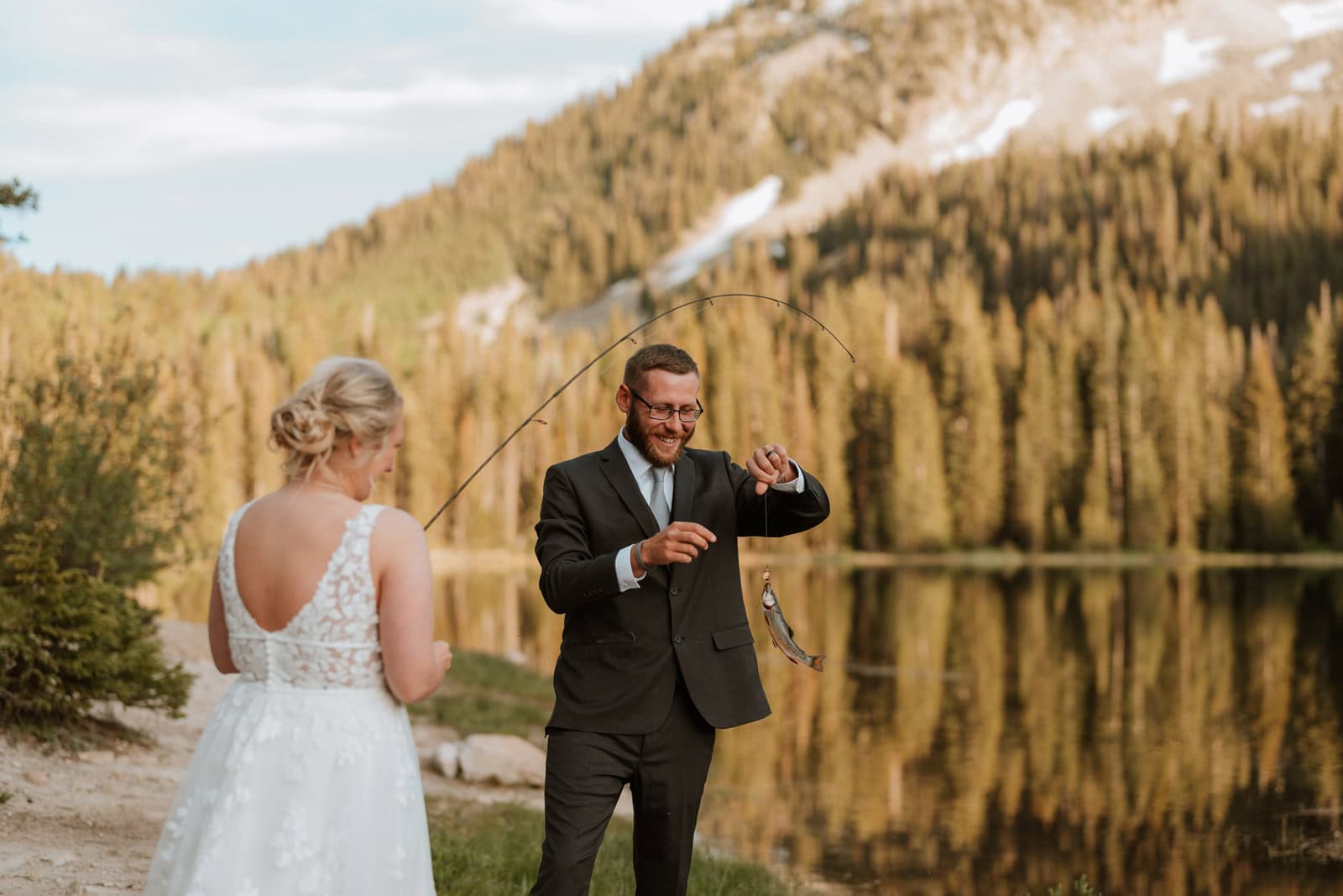 Couple fishing together by an alpine lake during an elopement in Colorado, celebrating with a caught fish and golden light reflecting off the water and trees.