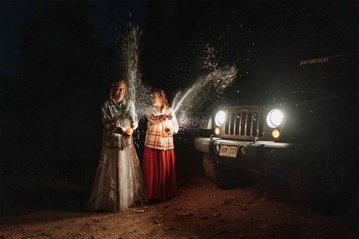 Two brides celebrate by popping champagne in the dark in front of their Jeep lights after eloping in Crested Butte.