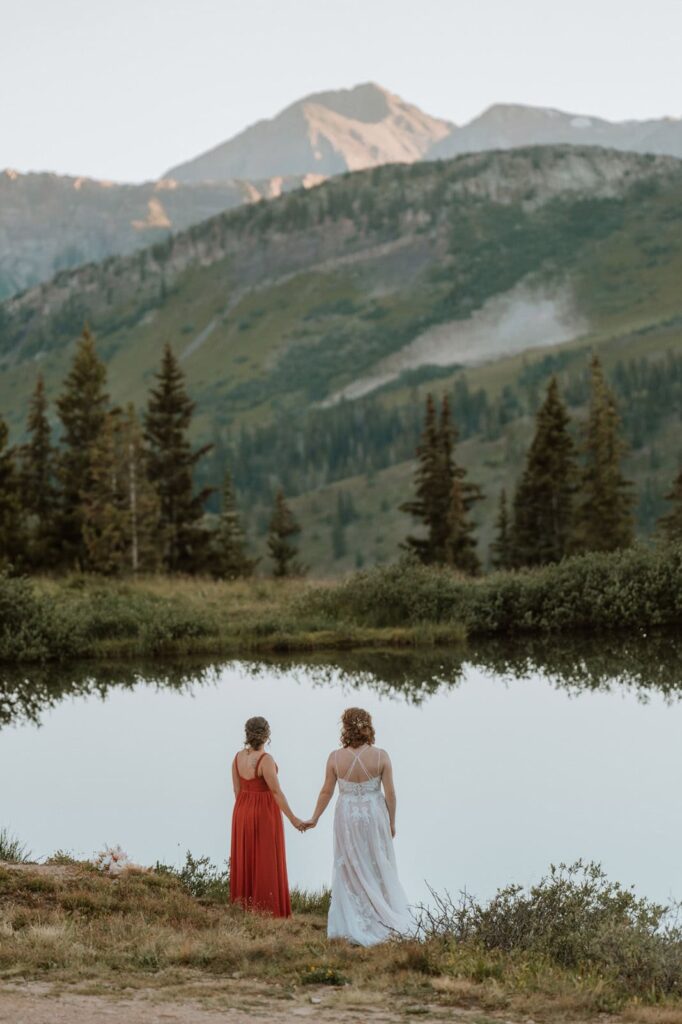 Two brides holding hands beside an alpine lake during a lesbian elopement in Crested Butte, surrounded by pine trees and mountain peaks reflected in the water.