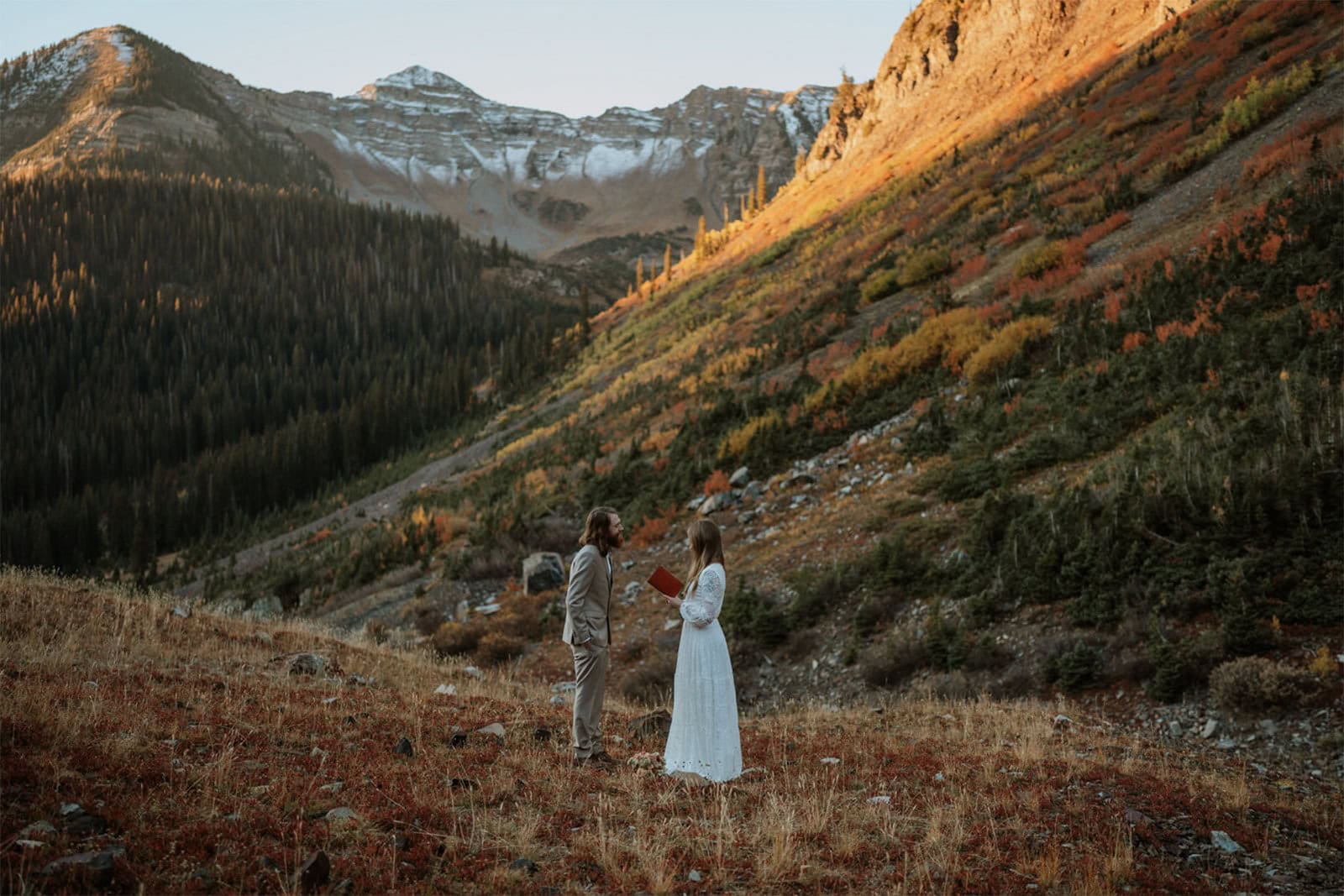 Bride and groom share their elopement vows at sunrise during their Crested Butte elopement. They are surrounded by snowcapped mountains and the sun is just starting to light them up. You can also see colorful fall foliage behind them.