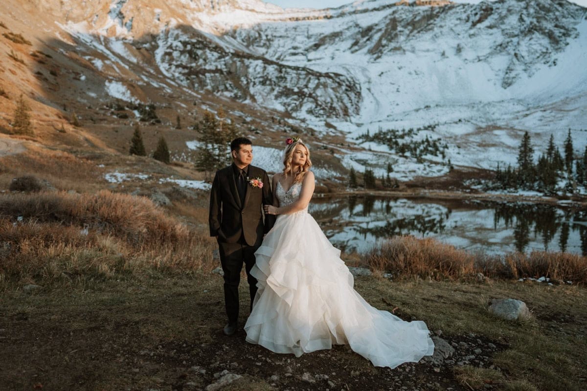 Bride holds onto her groom's arm and they both look off into the distance. The sunset is shining on their faces and the mountains behind them.