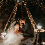 Bride and groom kiss while sitting under a tipi with wooden logs put together. The logs are wrapped in strightlights and the lanterns are lighting up the couple because it's dark outside for this night couples photo.