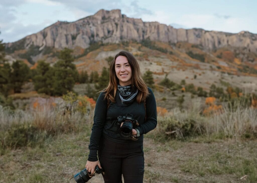 Colorado elopement photographer, Paige Weber, is smiling for the camera and holding two Sony cameras. There are Colorado mountains and fall colors in the background behind her.