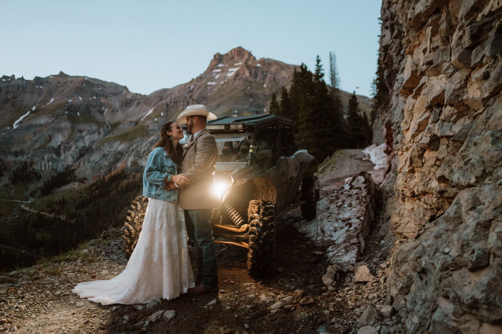 Couple in wedding attire standing together beside a 4x4 UTV on a narrow mountain road with rocky cliffs near Ouray.