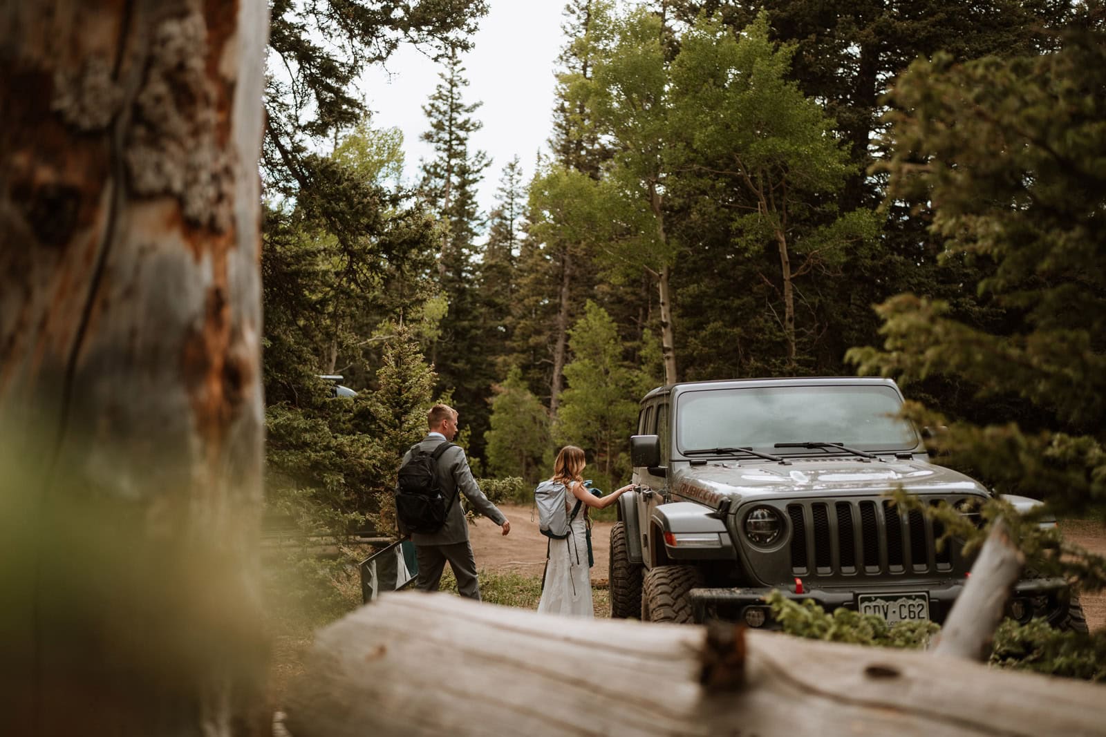 Couple in wedding attire walking toward a parked jeep along a forest road surrounded by pine trees during an outdoor wedding day in Colorado.