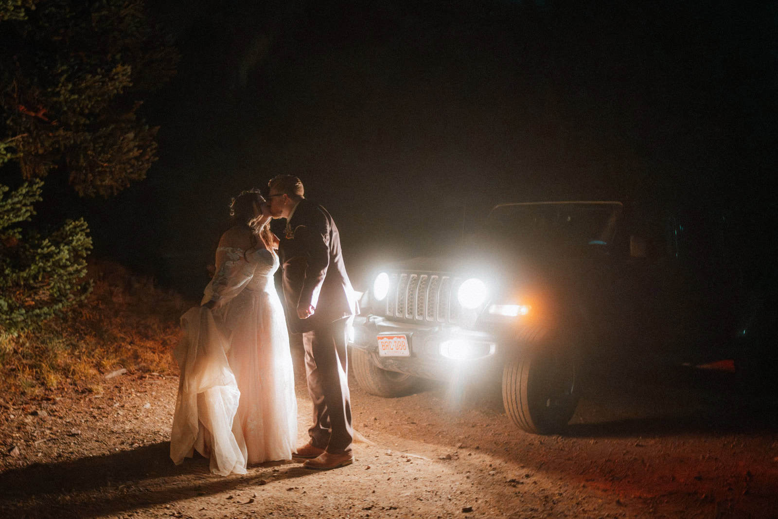 Couple in wedding attire standing close together on a dirt road at night, softly lit by the headlights of a parked jeep with surrounding darkness and trees.