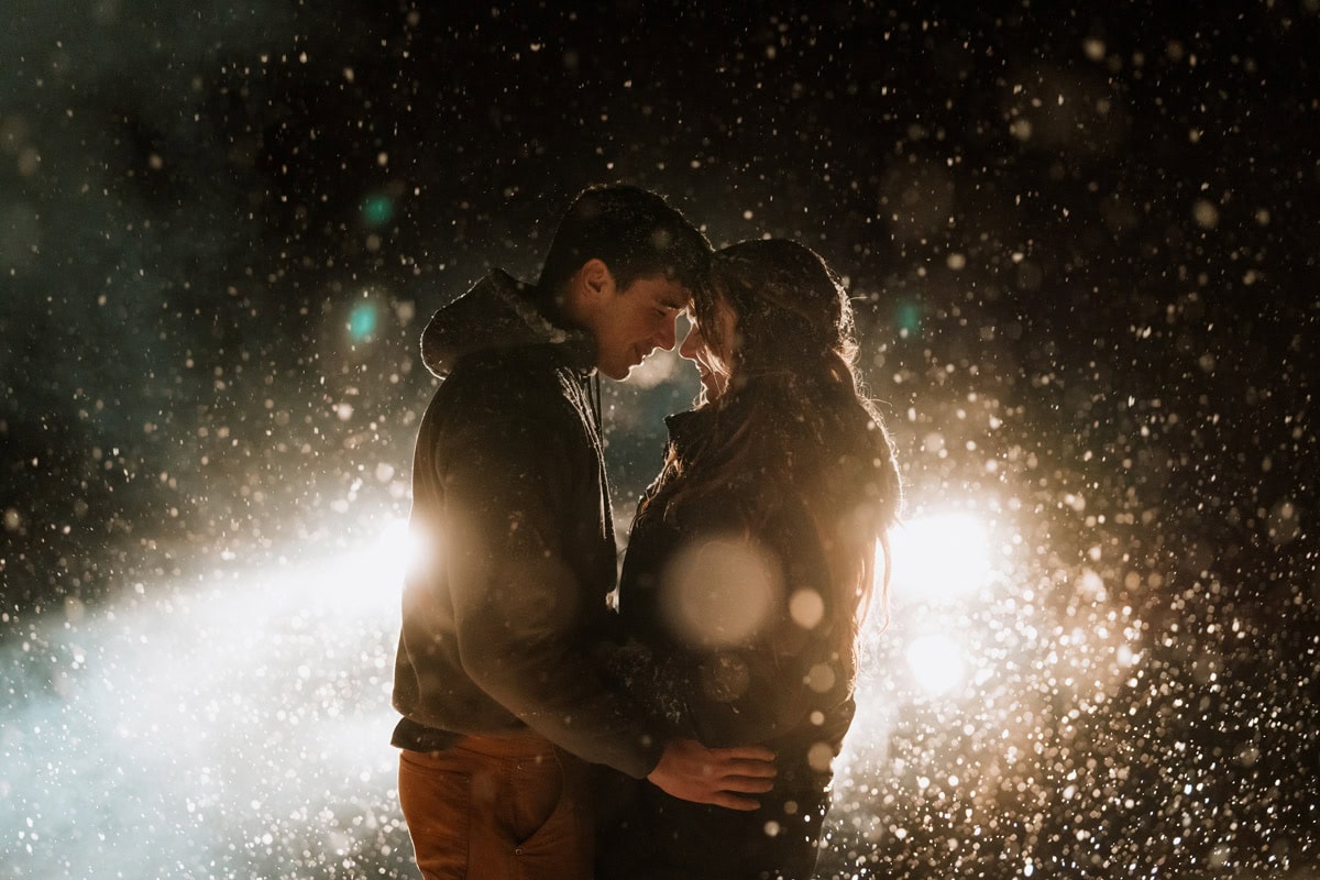 Couple embracing during a snowy nighttime winter elopement, softly backlit with falling snow all around them