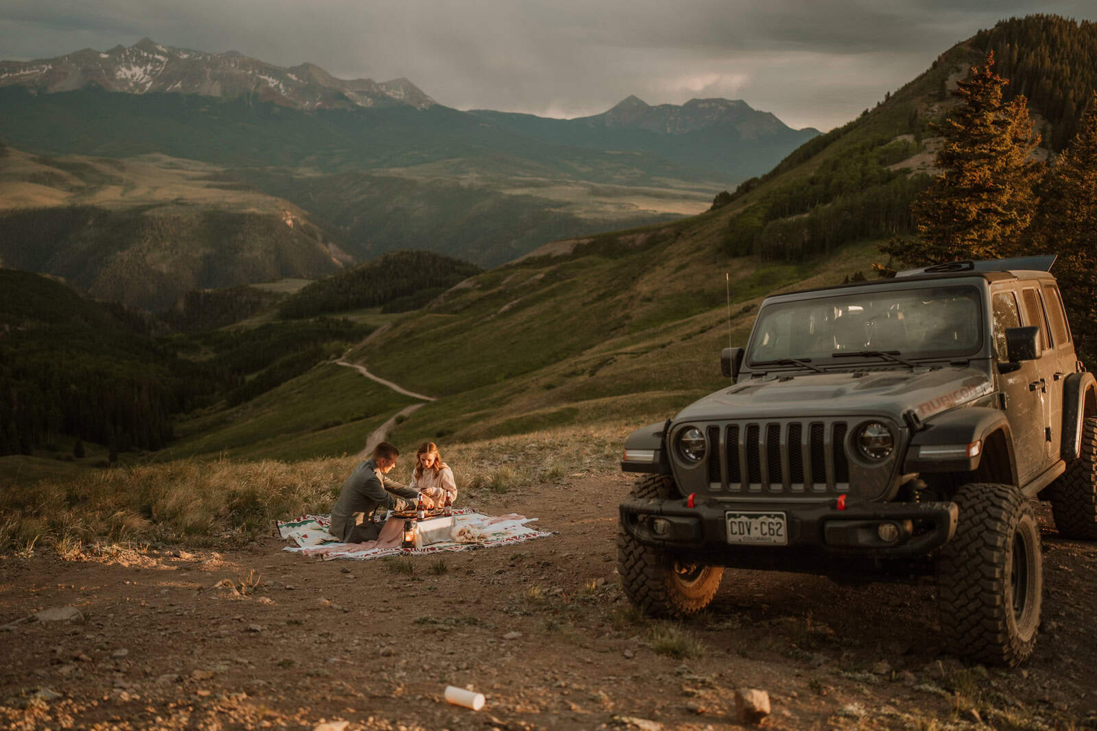 Couple sitting on a blanket sharing a picnic beside a parked jeep on a mountain overlook at sunset during a jeep wedding near Telluride.