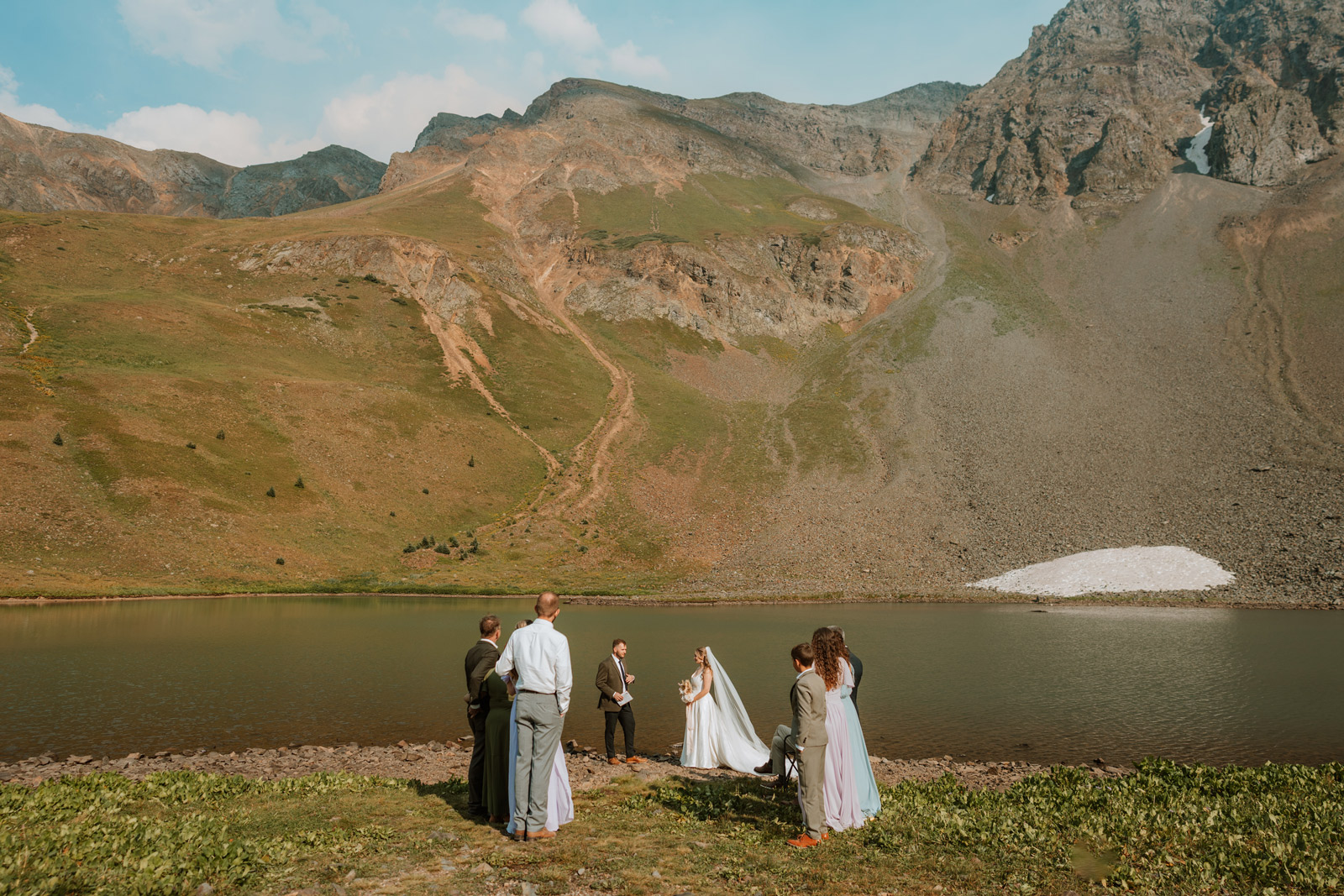 Bride and groom hold hands in front of a glacial alpine lake surrounded by dramatic rocky peaks during a small wedding in Colorado.