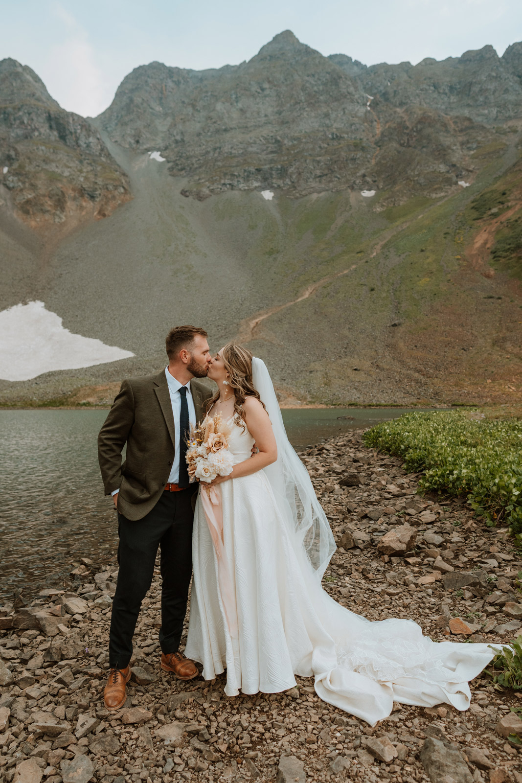 Bride and groom kiss on the rocky shoreline of an alpine lake in the backcountry during their San Juan Mountains elopement.
