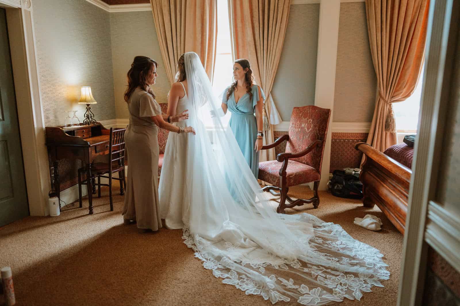 Two women help the bride with her veil in a sunlit window of the Beaumont Hotel, captured during the getting ready portion of the Ouray wedding day.